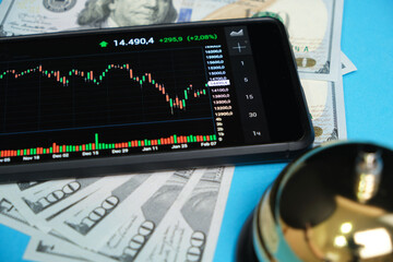 Stock exchange chart on smartphone screen, bell and hundred dollar bills blue background.Concept,...