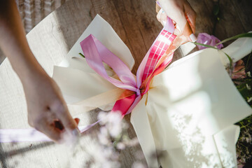 women's hands tie with a pink ribbon a bouquet of flowers, wrapped in paper, lying on a wooden table