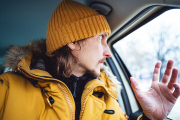 The man looks out the car window. Frozen car window in winter. A guy in bright clothes sits in the...