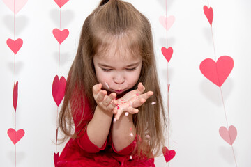 Concept Valentines Day. A girl child with long hair in a red jacket blows on a confetti in her palms.