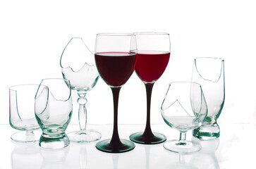 Two glasses with red wine surrounded by broken glasses on a white background.
