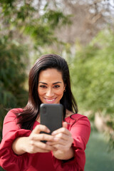 Charming Hispanic female smiling and taking self portrait while standing on street with lush green trees in city on summer day