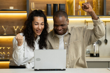 Mixed race woman and African American man get happy winning lottery. Cheerful multiracial couple looks at screen of laptop at table in kitchen close view