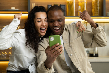 Young multiracial couple check results of competition in mobile phone. Mixed race woman and African American man get happy after win sitting in kitchen closeup