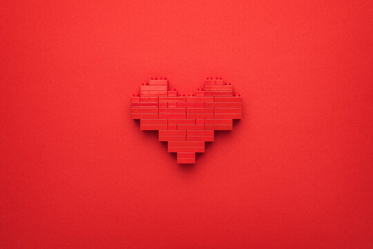 Red heart symbol made of plastic building blocks. Flat lay image of like button on red background. Top-down composition of toy pixelated heart model. Minimalist photo of stylized red love symbol.