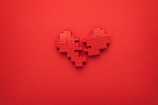 Photo of toy pixelated broken-down heart. Minimalist photo of stylized red relationship end symbol. Red broken heart made of building blocks. Flat lay image of shattered heart on red background.