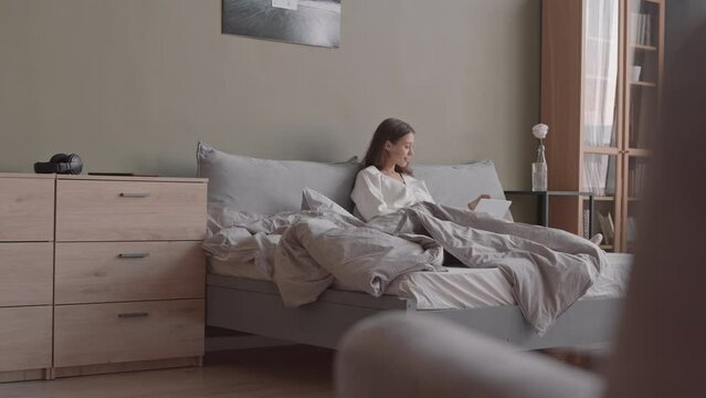 Slowmo shot of young Caucasian woman with laptop having breakfast in bed at cozy home