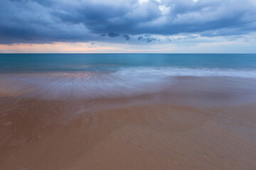 The view of the beach has white foam waves hitting the sand with turquoise water. Fluid dynamics, wave action in low light at sunset