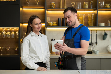 Foreman in uniform shows woman plan of kitchen renovation in tablet standing against countertop. Housewife uses services of professional specialist