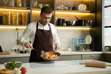 Young chef man in apron tosses steamed vegetables on pan above stove in kitchen. Cook prepares dish...