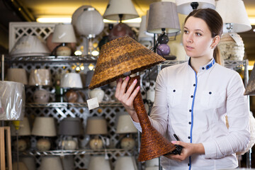 positive adult woman is buying lamp in the furniture store.