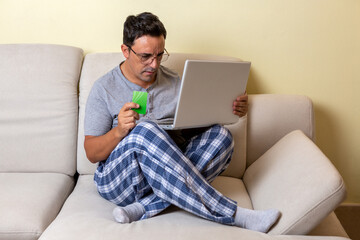 Young Caucasian male sitting on a couch using a laptop computer and credit card shopping online