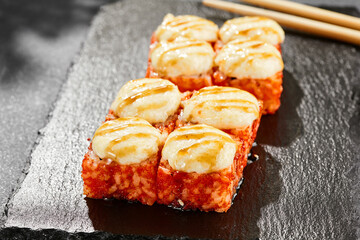 Baked Maki sushi on dark slate. Hot california maki with cheese. Sushi roll with tobiko outside, baked cheese topped. Style concept japanese menu with black background, leaves and hard shadow.
