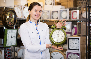 positive adult woman is holding clock in the furniture store.
