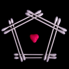 Cotton buds are arranged on a shiny black surface with the shape of a house and a red heart in the center