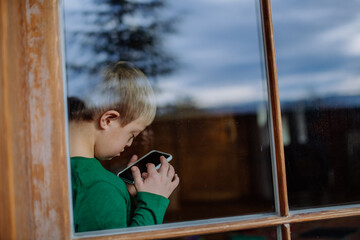 Obraz premium Little boy with Down syndrome using smartphone at home, shot through window.