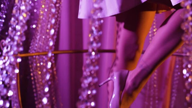 Elegant woman in silvery skirt in silvery high-heeled shoes lifts her slender leg in purple light. Crystal beads of large decorative chandelier