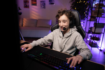 Young player plays computer at night in a room lit by led light, boy, teenager talks jokes laughs with teammates through headset, online sports championship, virtual world, winning round