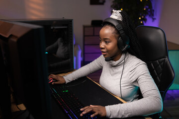 Fototapeta na wymiar Pro gamer wearing headphones with microphone talks to team members multiplayer game, girl with dark skin looks smiling contentedly into computer monitor, strategic thinking, shooters