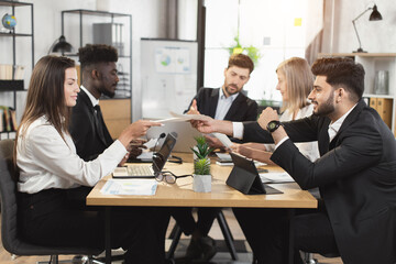 Multi ethnic business partners discussing common project during office briefing. Men and women in stylish formal clothes sitting at desk with modern technology.
