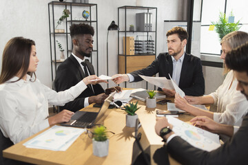 Multi ethnic team of competent financiers dressed in stylish formal clothes sitting together at office room and having brainstorming. Males and females colleagues discussing common business project.