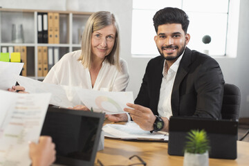 Young indian man and caucasian senior woman smiling and looking at camera during paperwork at office. Two company workers in formal wear cooperating together.
