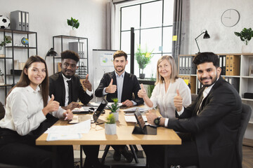 Team of five multiracial business people in formal clothes sitting together at office desk, smiling and looking at camera and showing thumb up. Conference meeting of diverse colleagues.