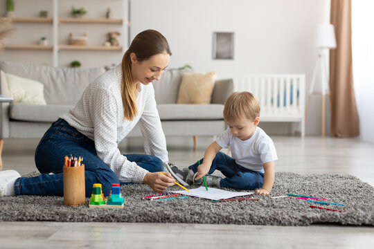 Leisure with baby. Happy mother and toddler son drawing with colorful pencils, relaxing together on floor in living room