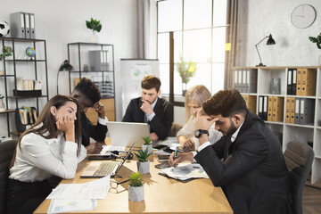 Group of pensive male and female office workers having urgent conference. Multi ethnic business colleagues sitting at desk and busy working on modern gadgets.