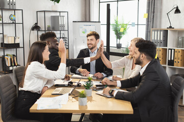 International team of business partners giving high five to each other during successful negotiation at boardroom. Men and women in formal clothes sitting at desk and smiling sincerely.