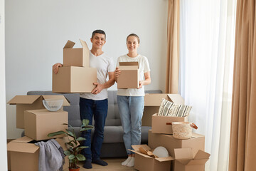 Full length portrait of woman and man, young couple wearing white t shirts standing with cardboard boxes, relocating to a new house, unpacking personal pile, looking smiling to camera.