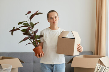 Portrait of sad upset woman wearing white t shirt standing with cardboard box, relocating to a new...