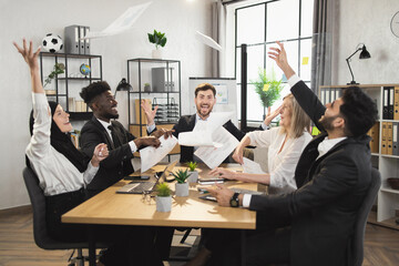 Team of happy diverse business partners sitting at desk and throwing up documents during successful meeting at office. Agreement, cooperation and people concept.