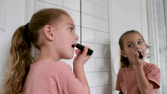 Close up young blonde little girl looks into it and useing chapstick lipstick at home mirror