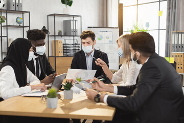 Multi ethnic financial specialists in formal wear and medical face masks gathering at boardroom for conference meeting. Brainstorming of male and female business people.