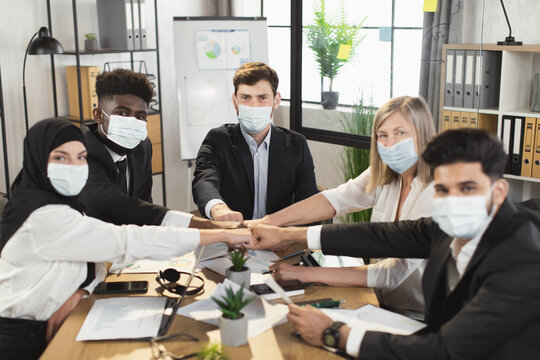 Group of multiracial business people in medical face masks stacking their fists in circle while sitting at conference room and looking at camera. People, work and pandemic concept.