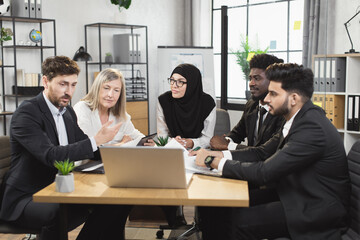 Team of male and female business partners using modern laptop for video call at boardroom. Colleagues in formal clothes sitting together at desk, talking and gesturing.