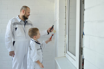 Naklejka na ściany i meble Happy father and son dressed in identical white jumpsuits painting a wall in their home.