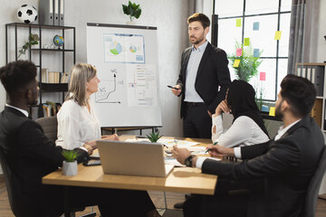 Successful man in black suit standing near flip chart and talking with multicultural colleagues during conference meeting. Group of business people gathering at office room for briefing.