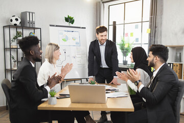 Group of multiracial business people in formal wear sitting at office desk and clapping hands after successful speech of their caucasian male colleague. Briefing of competent financiers.