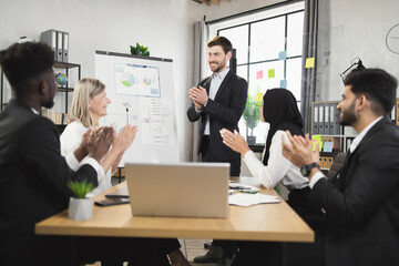 Positive of multicultural business partners in formal wear sitting at boardroom and clapping in hands. Happy colleagues satisfied with successful meeting at modern office.