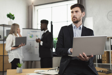 Caucasian man in suit using laptop and looking aside while leaning on desk at modern office. Multiracial company workers standing on background and talking.
