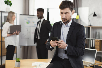 Caucasian businessman in suit using modern smartphone and looking at camera while sitting at office. Blur background of multiracial partner talking near flip chart.