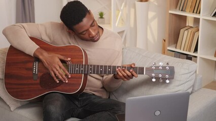 Guitar course. Internet music class. Creative hobby. Diligent man guitarist learning string instrument chords lesson online at laptop on couch at home.