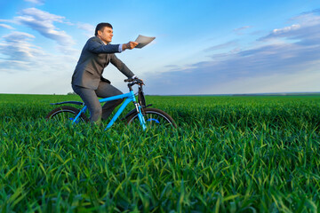 businessman dressed in a business suit, rides a bicycle through a green grass field, he has a briefcase and documents, beautiful nature in spring, business concept