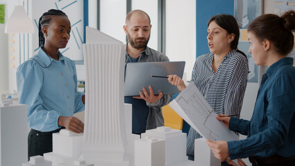 Colleagues working with laptop and blueprints to analyze building model in office. Workmates designing urban constructionwith laptop and maquette, doing teamwork for development.