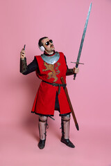 Studio shot of man, medieval warrior or knight listening to music in headphones with delightful expression isolated over pink background