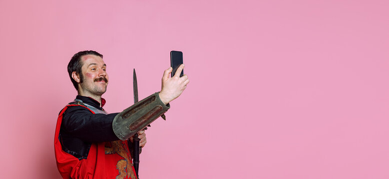 Comic portrait of medieval warrior or knight wearing wearing armor clothing using phone isolated over pink background. Comparison of eras, history