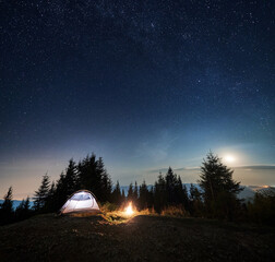 Illuminated tourist tent setting on grassy top of rocky mountain hill. Small evening campsite with tent and campfire on background of forest, silhouettes of mountains under starry colorful sky.