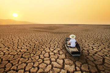 Young man paddle a boat at dry cracked earth with orange sky and hot weather of the sun. Metaphor Climate change, Drought and water crisis concept. - obrazy, fototapety, plakaty